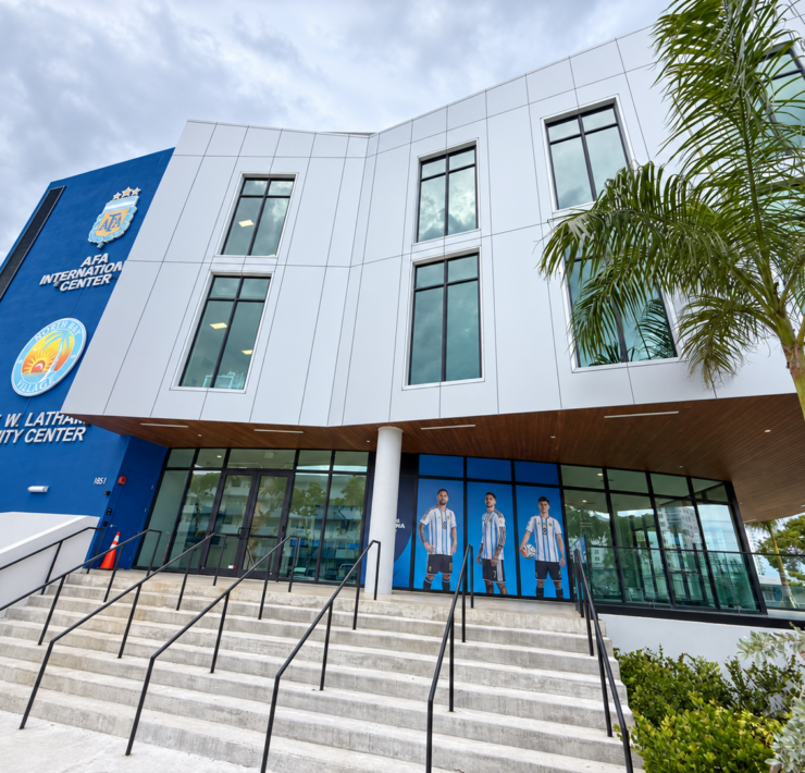 Stairs and railings leading to the glass entrance of the Brent W. Latham Community Center, with Argentina soccer player graphics and palm trees.