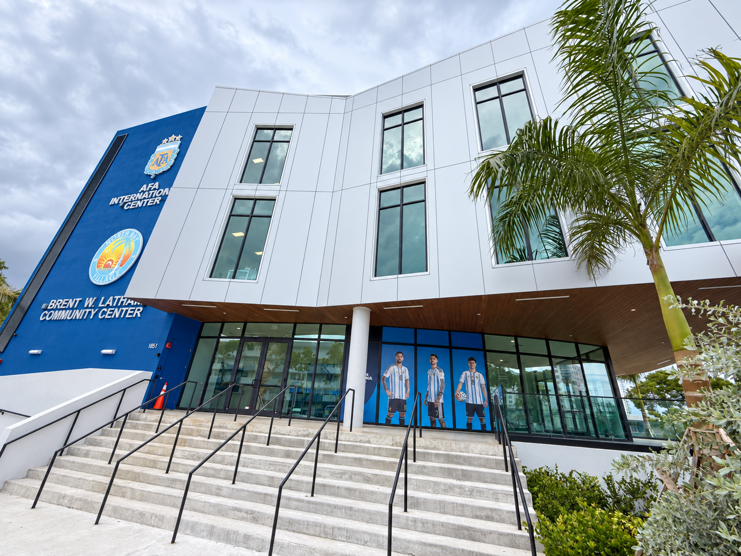 Stairs and railings leading to the glass entrance of the Brent W. Latham Community Center, with Argentina soccer player graphics and palm trees.