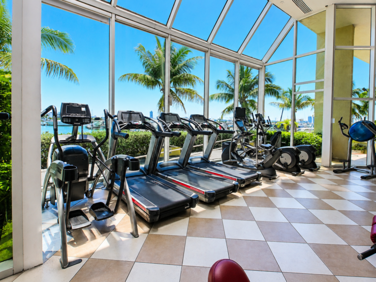 Cardio equipment at Treasure Drive Fitness in North Bay Village, beside large windows overlooking palm trees and the marina.